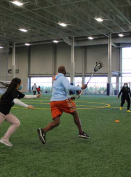 Man runs on indoor turf field with lacrosse stick, girl chases.